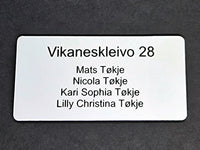 Bilde av hvitt postkasseskilt med navn og adresse gravert i svart tekst.